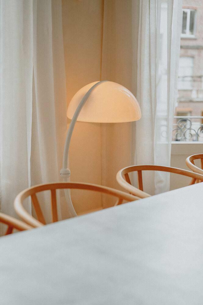 Close-up van inox eettafel en wishbone stoelen, met een sculpturale designlamp en zachte gordijnen in de achtergrond.